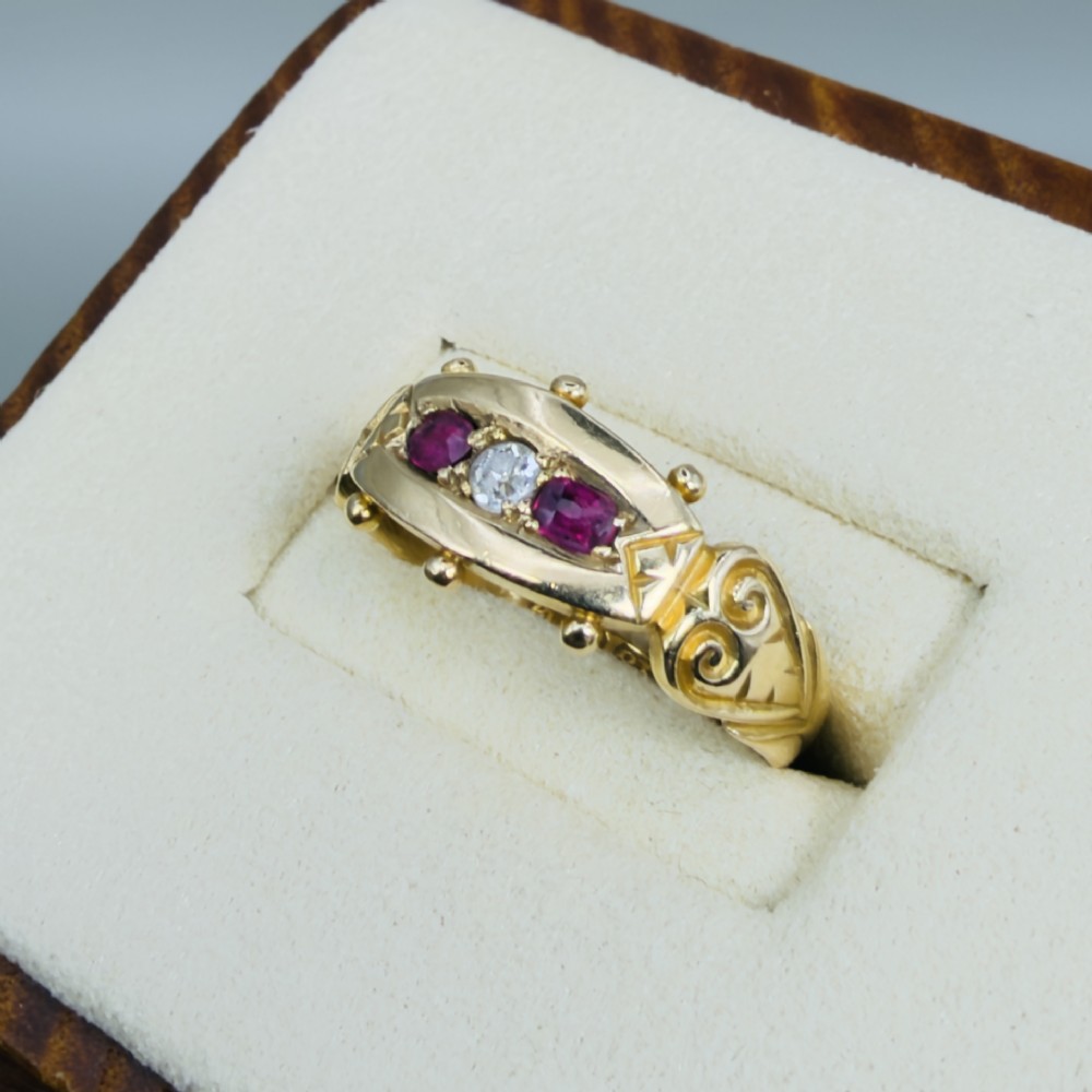 An 18ct yellow gold Edwardian trilogy ring set with a central round diamond flanked by two oval red rubies, featuring scroll detailing on the shoulders. View III