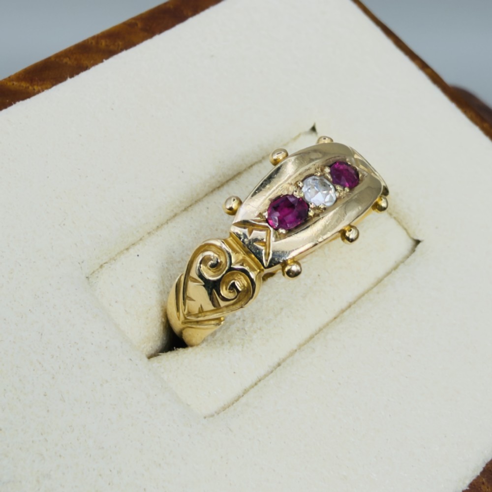 An 18ct yellow gold Edwardian trilogy ring set with a central round diamond flanked by two oval red rubies, featuring scroll detailing on the shoulders. View IV