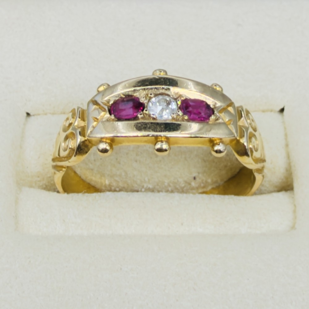 An 18ct yellow gold Edwardian trilogy ring set with a central round diamond flanked by two oval red rubies, featuring scroll detailing on the shoulders. View V