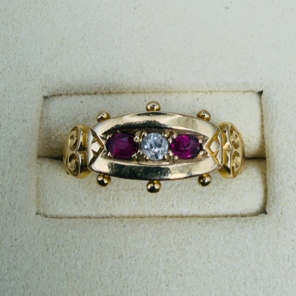An 18ct yellow gold Edwardian trilogy ring set with a central round diamond flanked by two oval red rubies, featuring scroll detailing on the shoulders. View VI
