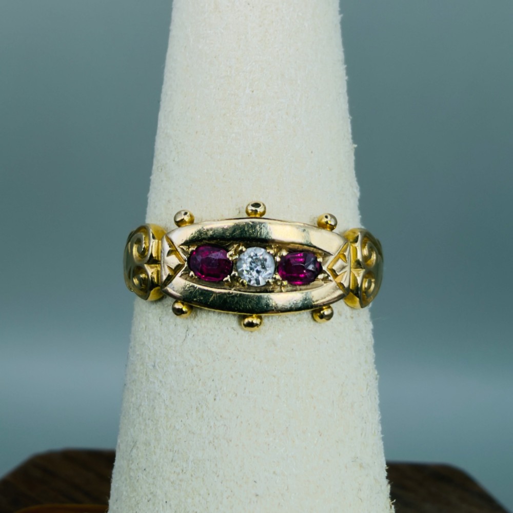 An 18ct yellow gold Edwardian trilogy ring set with a central round diamond flanked by two oval red rubies, featuring scroll detailing on the shoulders. View VII