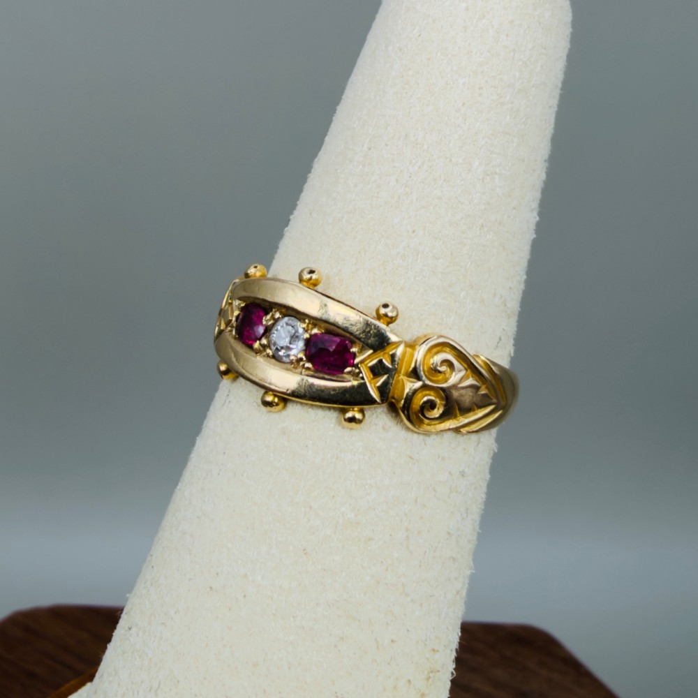 An 18ct yellow gold Edwardian trilogy ring set with a central round diamond flanked by two oval red rubies, featuring scroll detailing on the shoulders. View VIII