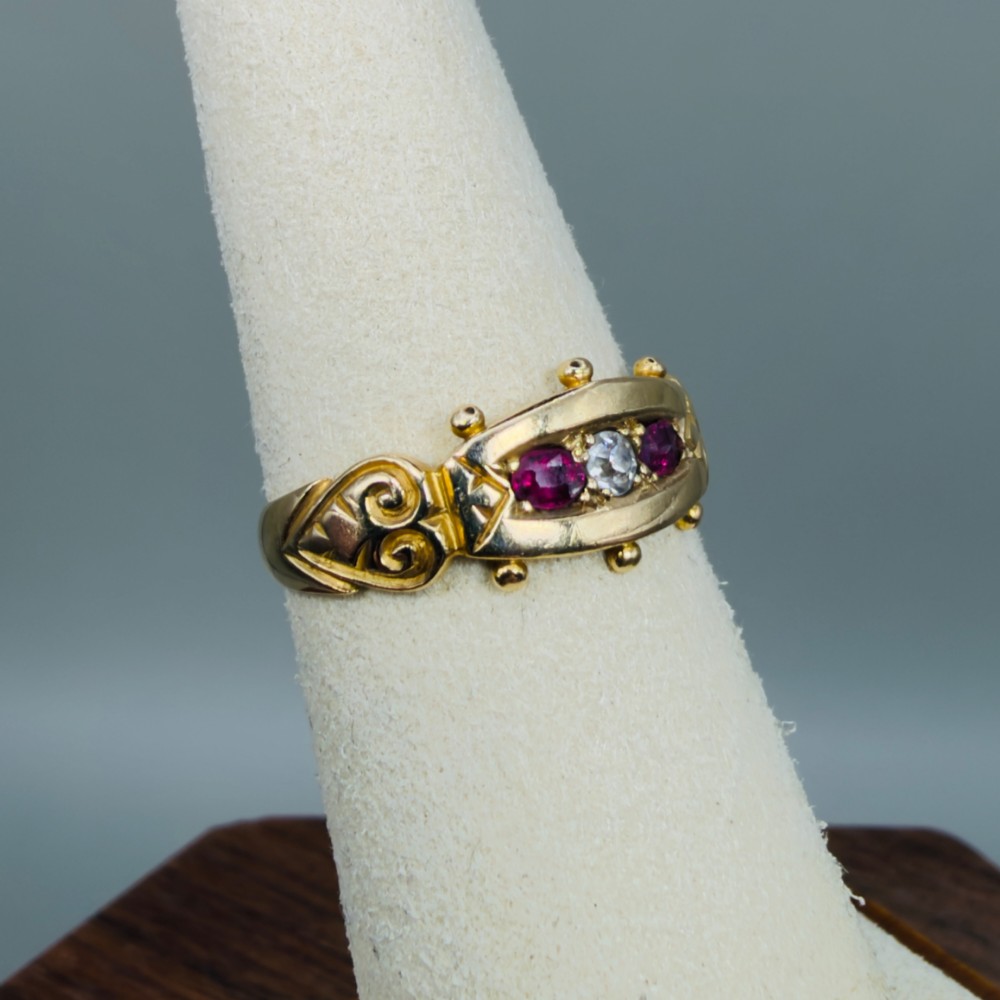 An 18ct yellow gold Edwardian trilogy ring set with a central round diamond flanked by two oval red rubies, featuring scroll detailing on the shoulders. View IX