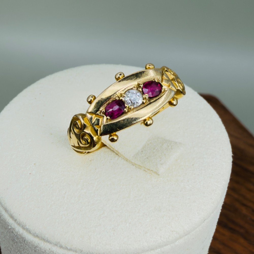 An 18ct yellow gold Edwardian trilogy ring set with a central round diamond flanked by two oval red rubies, featuring scroll detailing on the shoulders. View XI