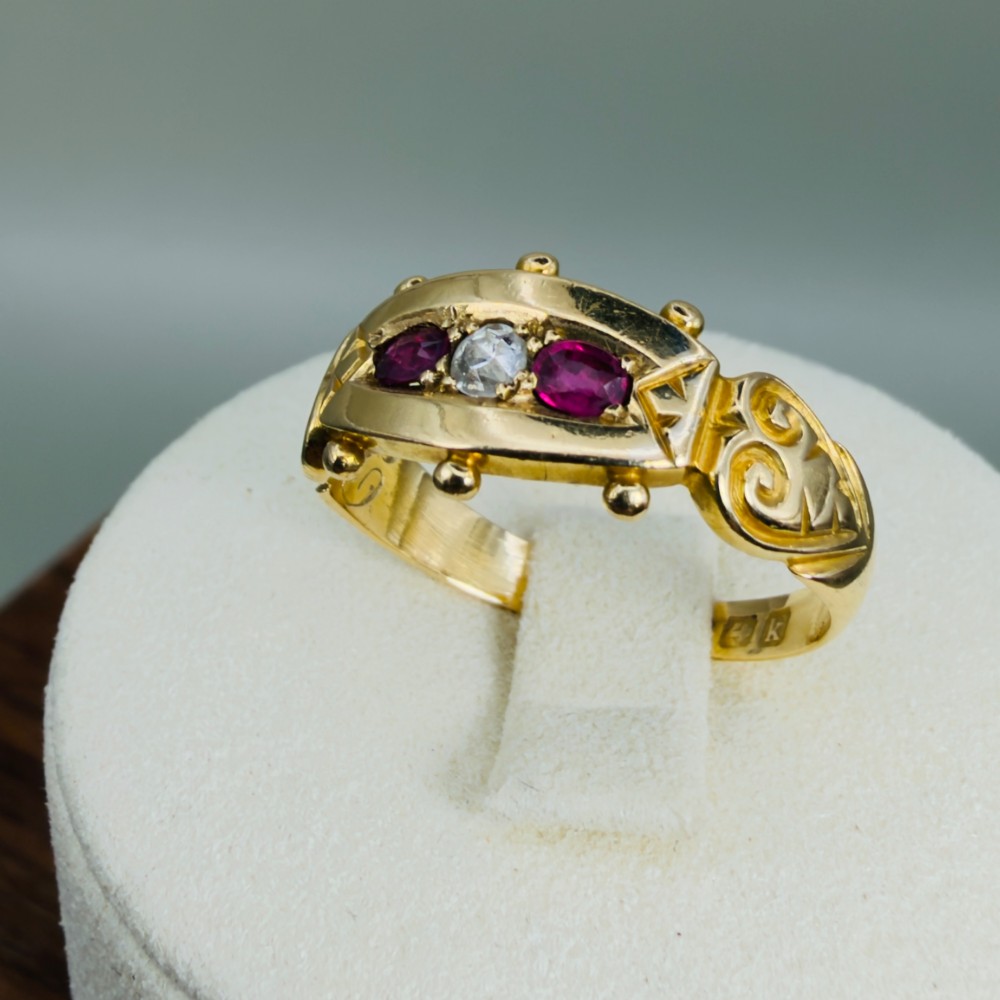 An 18ct yellow gold Edwardian trilogy ring set with a central round diamond flanked by two oval red rubies, featuring scroll detailing on the shoulders. View XII