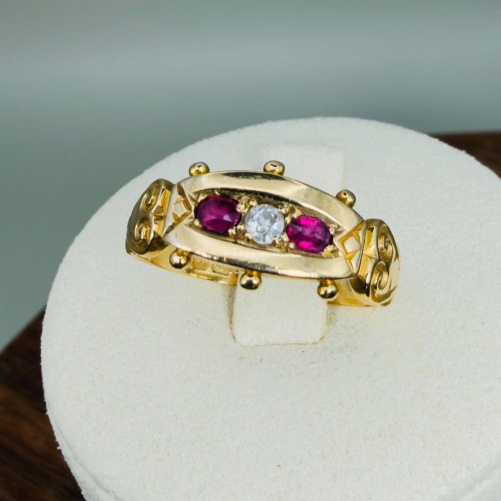 An 18ct yellow gold Edwardian trilogy ring set with a central round diamond flanked by two oval red rubies, featuring scroll detailing on the shoulders.