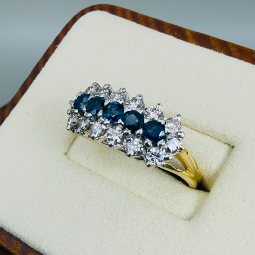 An 18ct gold cluster boat ring featuring five round blue sapphires surrounded by a halo of brilliant diamonds. View III