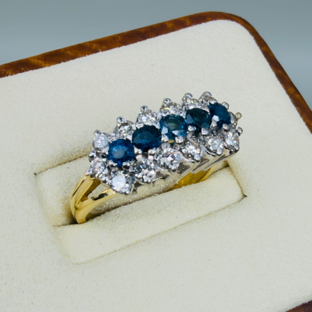 An 18ct gold cluster boat ring featuring five round blue sapphires surrounded by a halo of brilliant diamonds. View IV