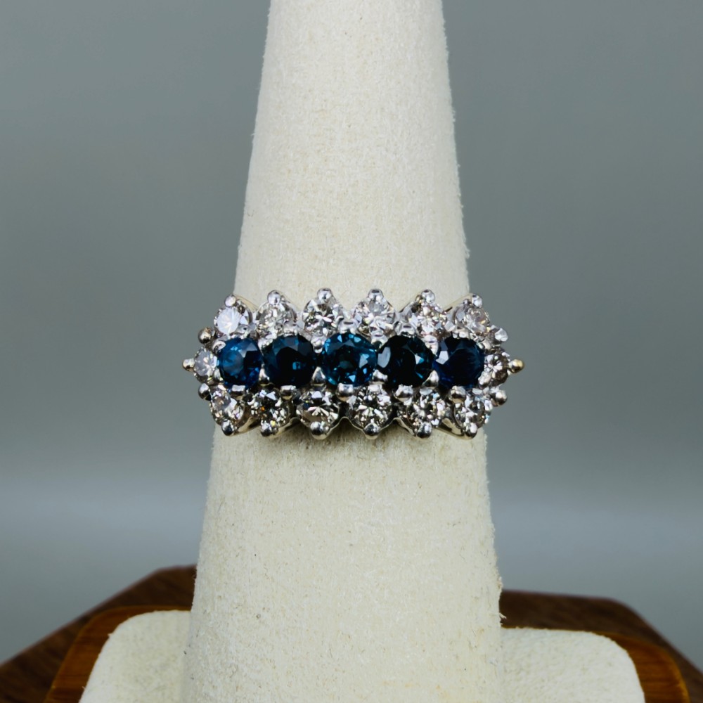 An 18ct gold cluster boat ring featuring five round blue sapphires surrounded by a halo of brilliant diamonds. View VI