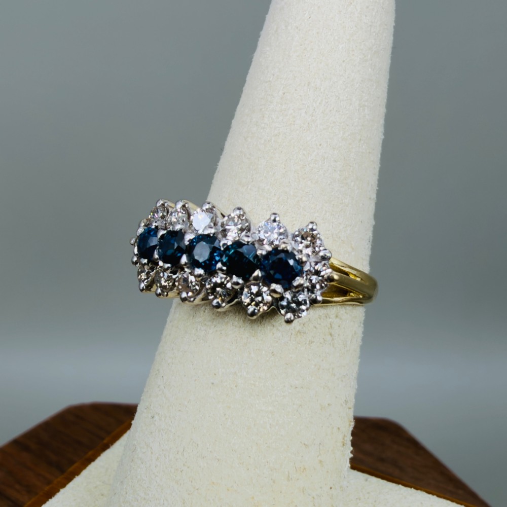 An 18ct gold cluster boat ring featuring five round blue sapphires surrounded by a halo of brilliant diamonds. View VII