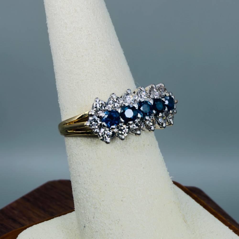 An 18ct gold cluster boat ring featuring five round blue sapphires surrounded by a halo of brilliant diamonds. View VIII