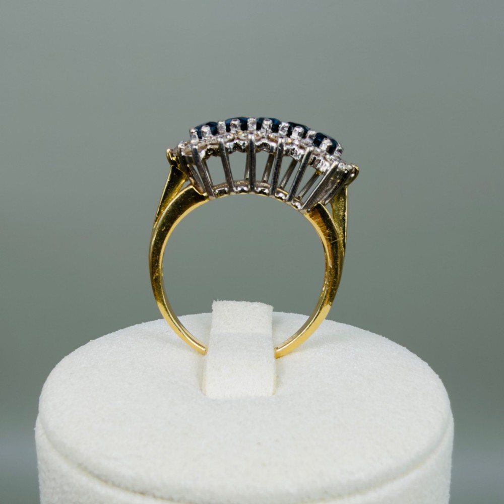 An 18ct gold cluster boat ring featuring five round blue sapphires surrounded by a halo of brilliant diamonds. View IX