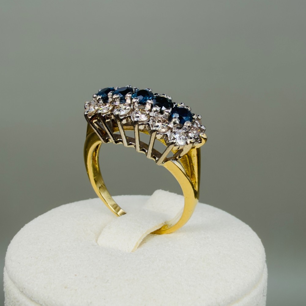 An 18ct gold cluster boat ring featuring five round blue sapphires surrounded by a halo of brilliant diamonds. View X