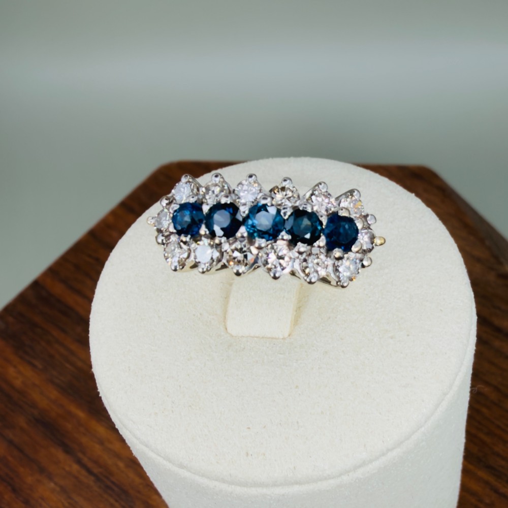 An 18ct gold cluster boat ring featuring five round blue sapphires surrounded by a halo of brilliant diamonds. View XI