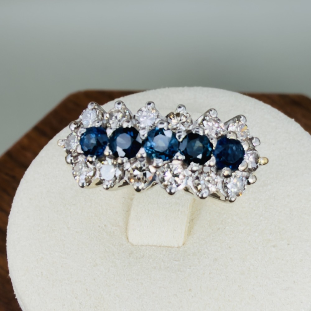 An 18ct gold cluster boat ring featuring five round blue sapphires surrounded by a halo of brilliant diamonds.