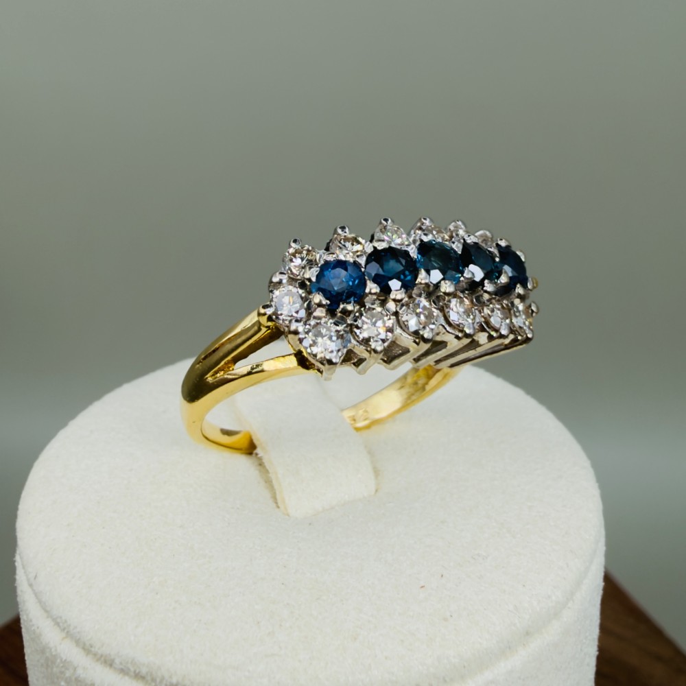 An 18ct gold cluster boat ring featuring five round blue sapphires surrounded by a halo of brilliant diamonds. View XII