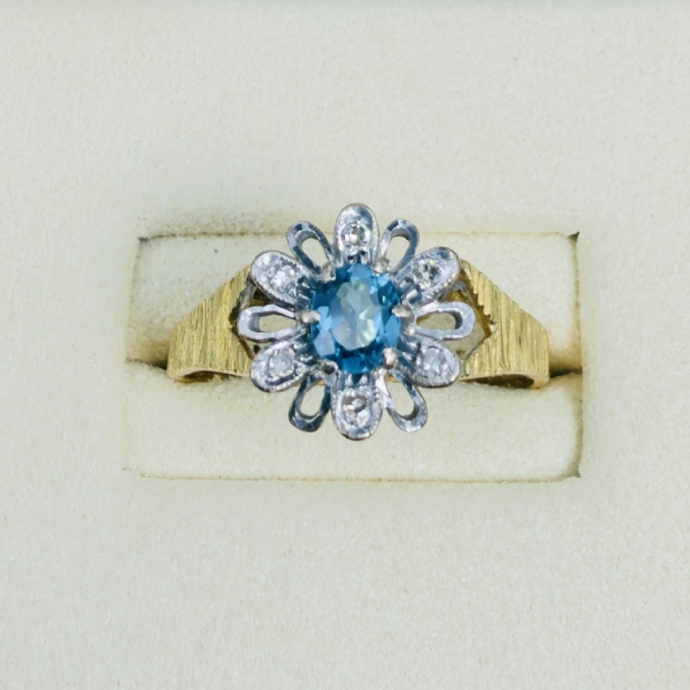 An 18ct yellow gold ring featuring a central oval blue aquamarine framed by an openwork floral halo set with diamonds and textured shoulders.
