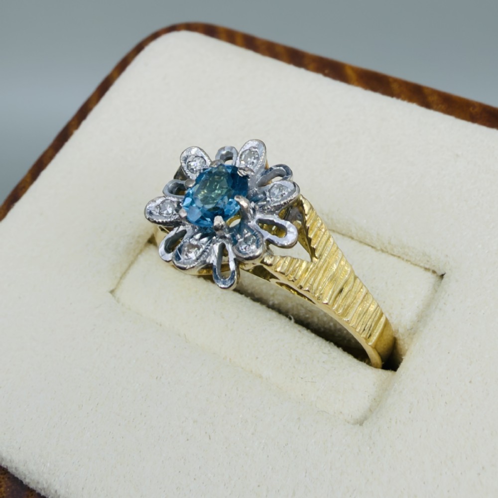 An 18ct yellow gold ring featuring a central oval blue aquamarine framed by an openwork floral halo set with diamonds and textured shoulders. View II