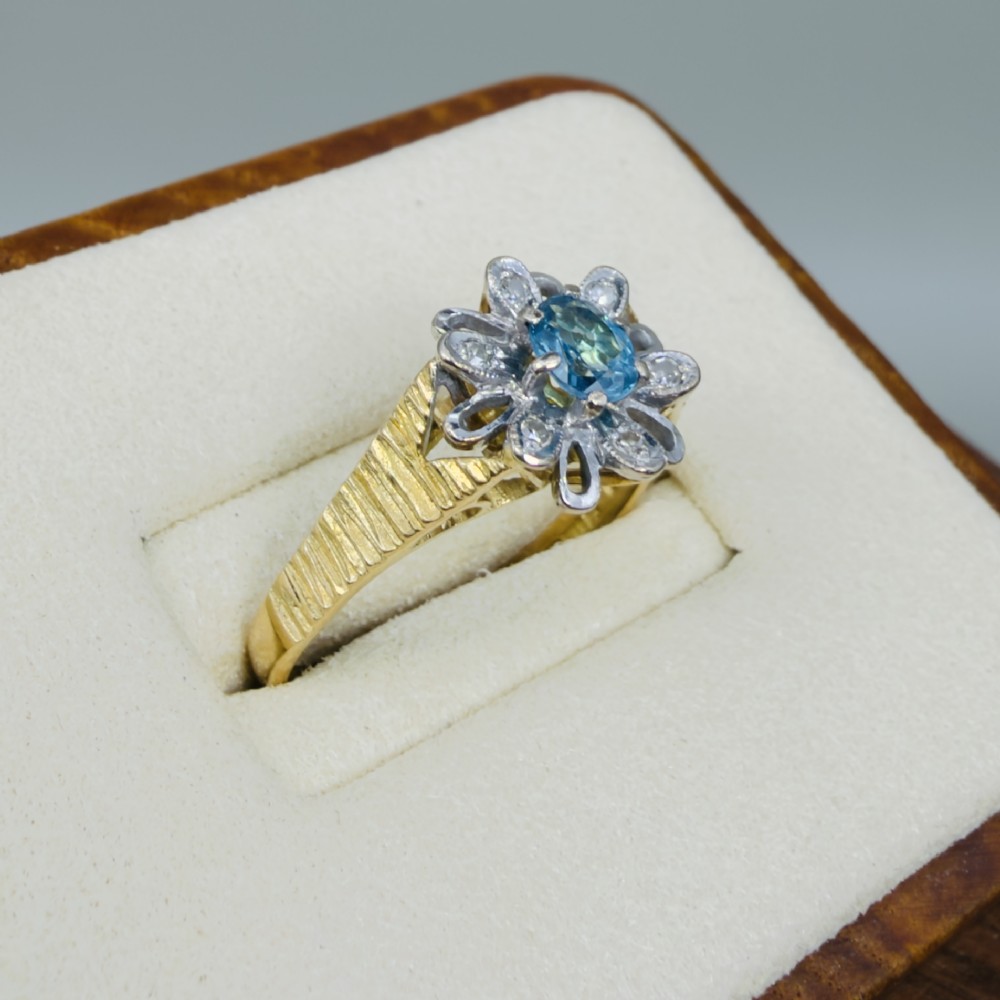 An 18ct yellow gold ring featuring a central oval blue aquamarine framed by an openwork floral halo set with diamonds and textured shoulders. View III