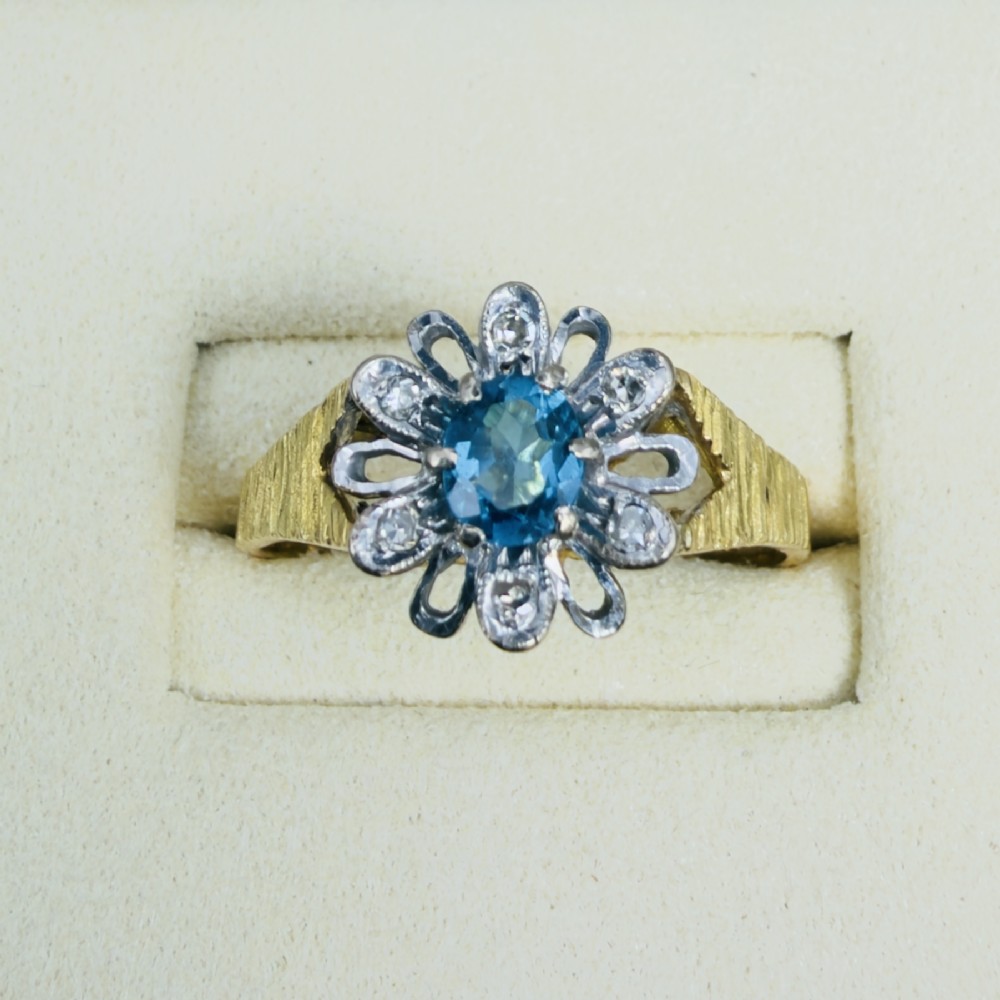 An 18ct yellow gold ring featuring a central oval blue aquamarine framed by an openwork floral halo set with diamonds and textured shoulders. View IV
