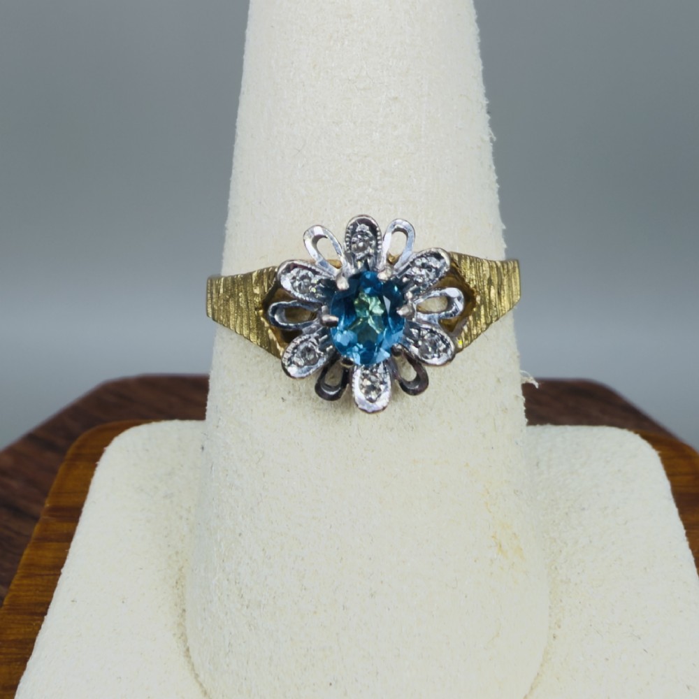 An 18ct yellow gold ring featuring a central oval blue aquamarine framed by an openwork floral halo set with diamonds and textured shoulders. View V