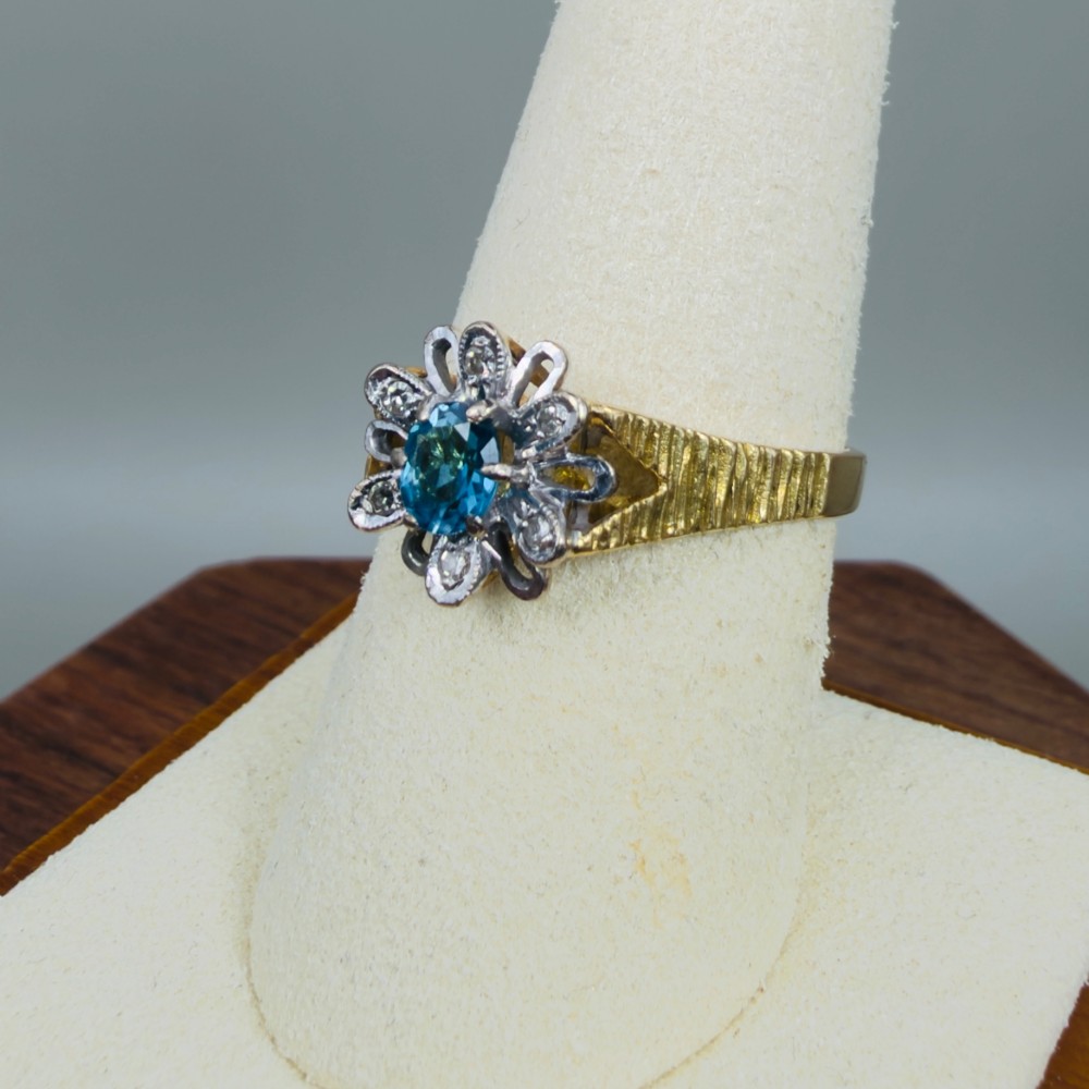 An 18ct yellow gold ring featuring a central oval blue aquamarine framed by an openwork floral halo set with diamonds and textured shoulders. View VI