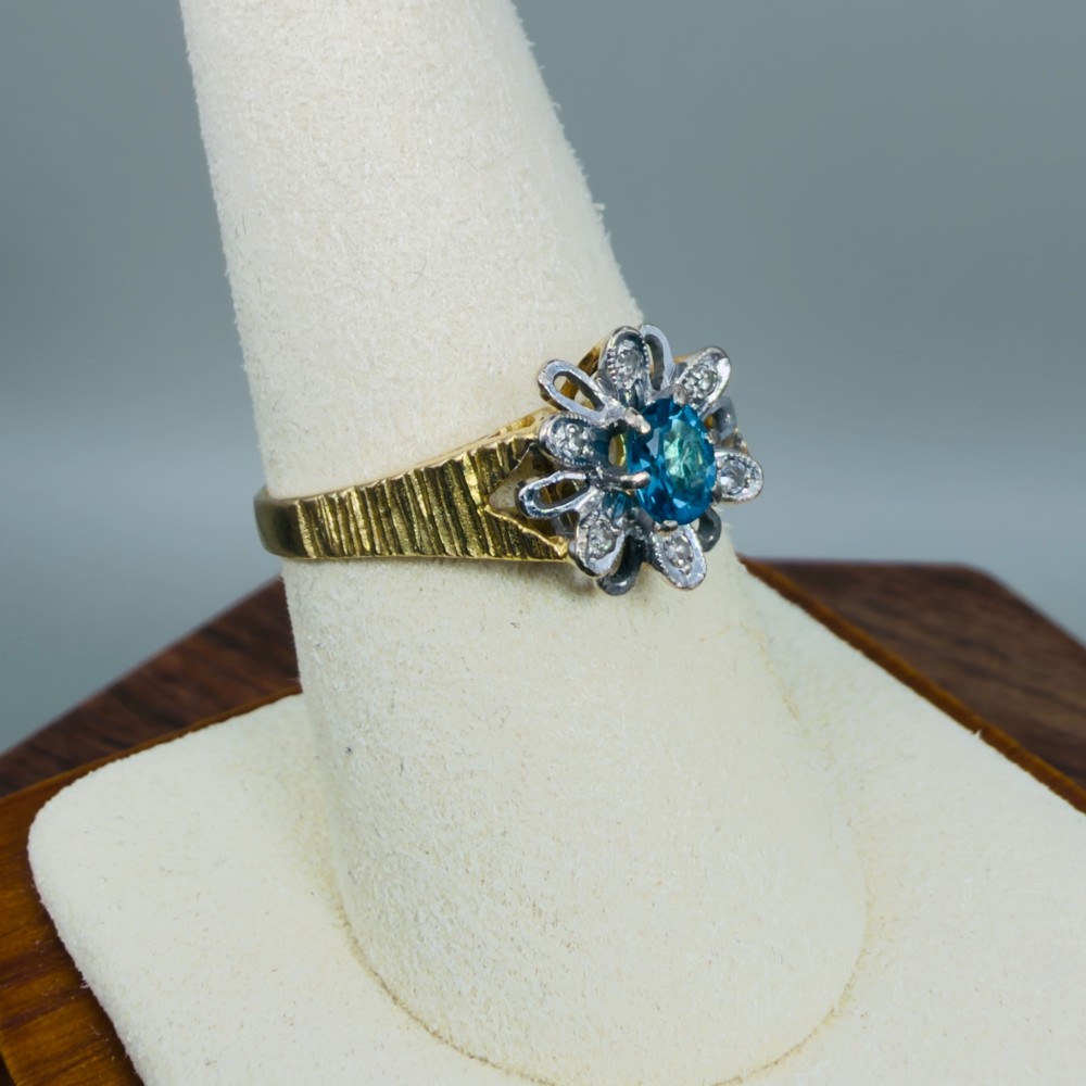 An 18ct yellow gold ring featuring a central oval blue aquamarine framed by an openwork floral halo set with diamonds and textured shoulders. View VII