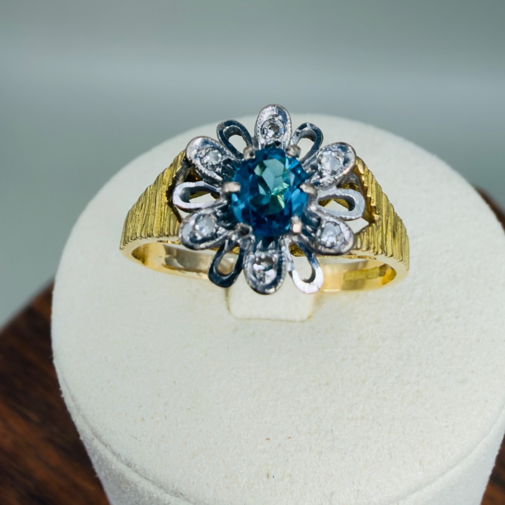 An 18ct yellow gold ring featuring a central oval blue aquamarine framed by an openwork floral halo set with diamonds and textured shoulders. View VIII