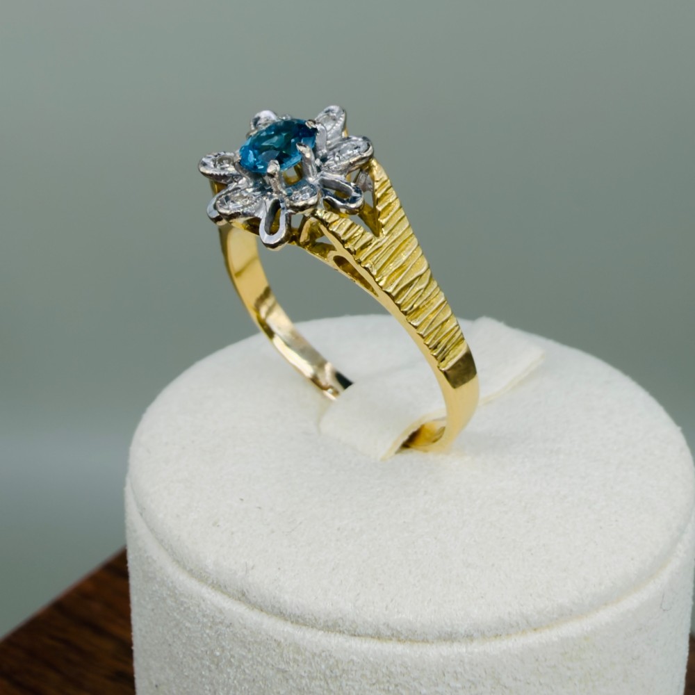 An 18ct yellow gold ring featuring a central oval blue aquamarine framed by an openwork floral halo set with diamonds and textured shoulders. View IX