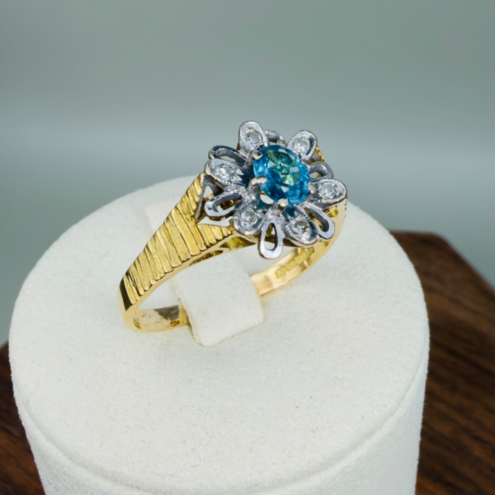 An 18ct yellow gold ring featuring a central oval blue aquamarine framed by an openwork floral halo set with diamonds and textured shoulders. View X