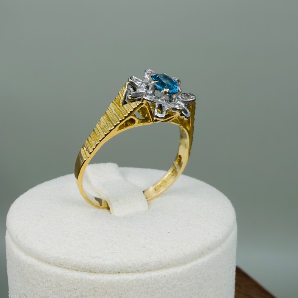 An 18ct yellow gold ring featuring a central oval blue aquamarine framed by an openwork floral halo set with diamonds and textured shoulders. View XI