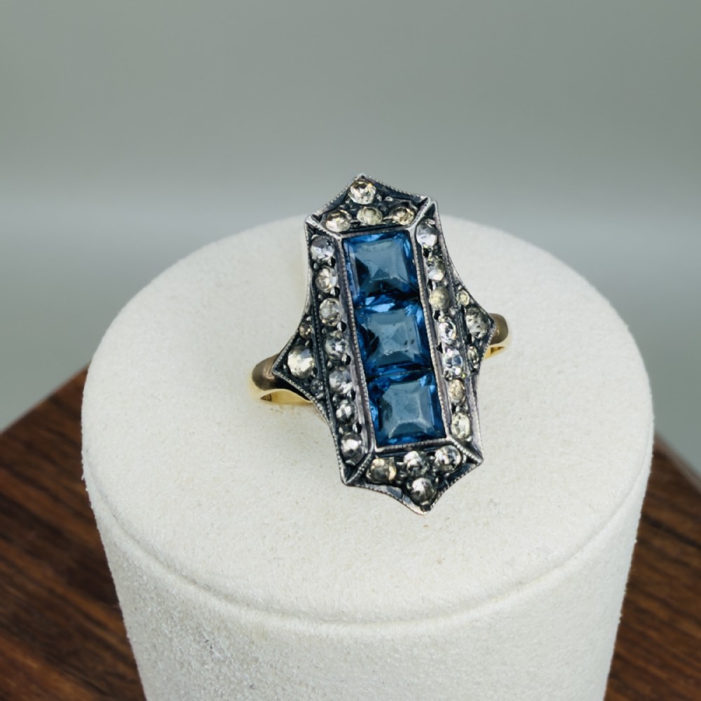 An Art Deco 18ct gold ring featuring a central vertical row of three square blue stones framed by white paste stones in an elongated geometric silver setting.