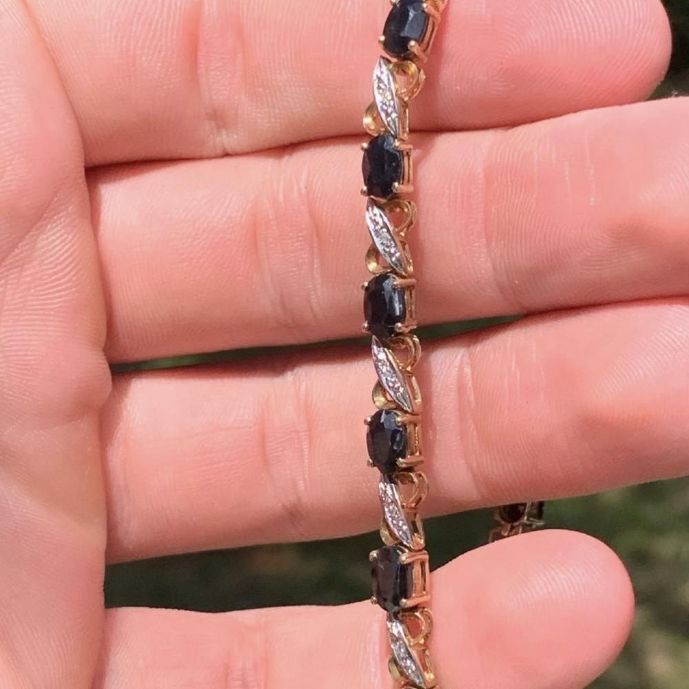 A 9ct yellow gold link bracelet set with oval-cut dark blue sapphires and accent diamond links, worn on a wrist. View V