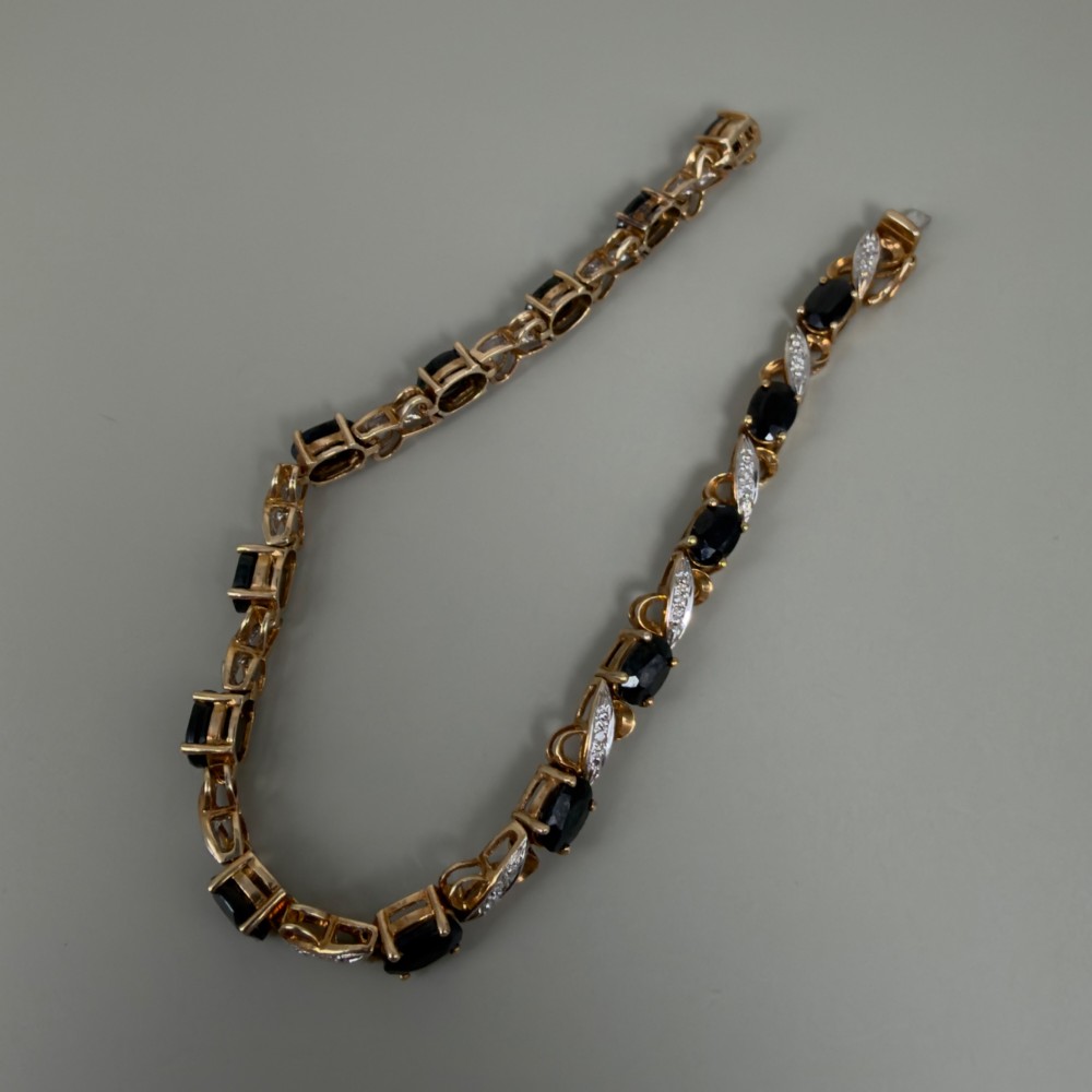 A 9ct yellow gold link bracelet set with oval-cut dark blue sapphires and accent diamond links, worn on a wrist. View X