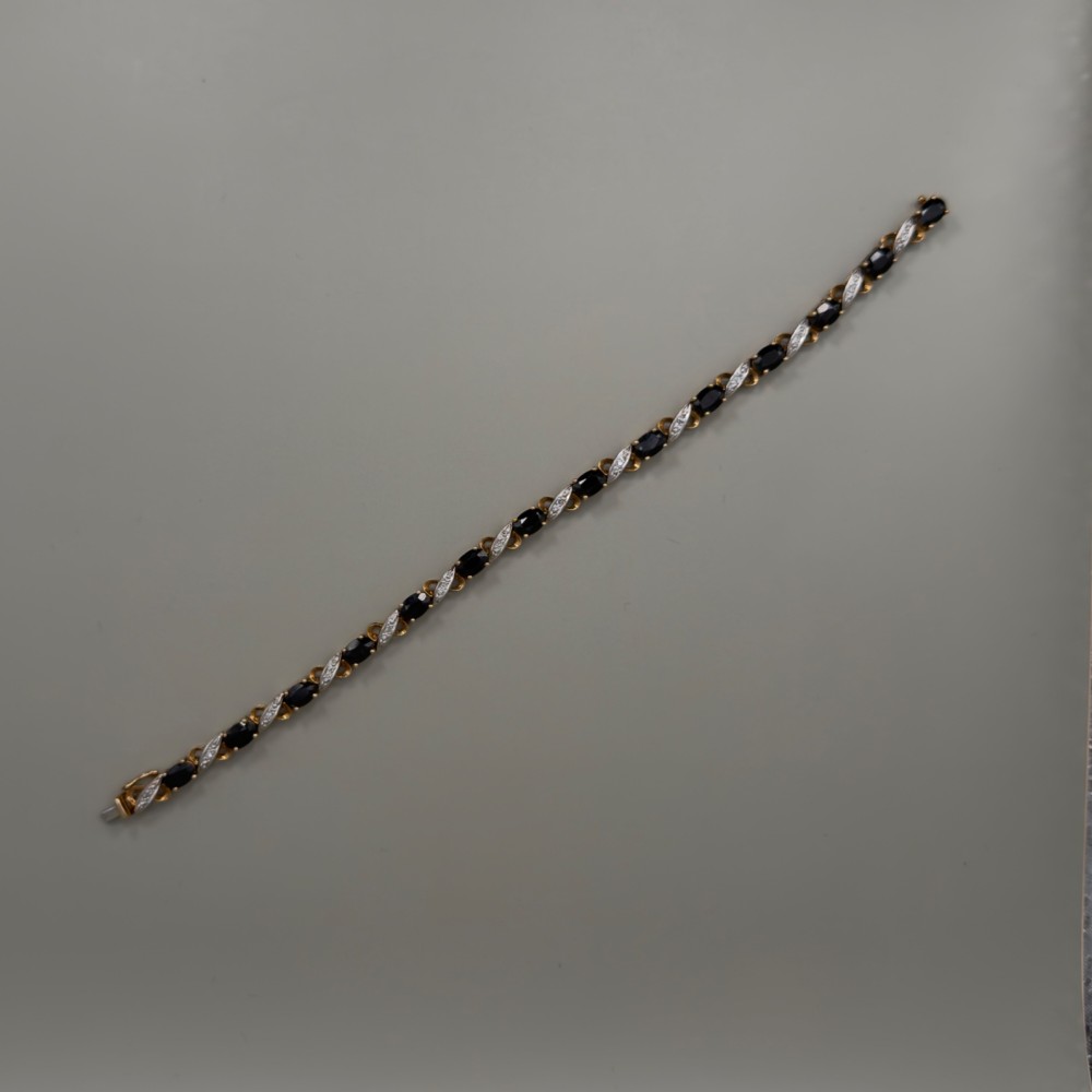 A 9ct yellow gold link bracelet set with oval-cut dark blue sapphires and accent diamond links, worn on a wrist. View XI