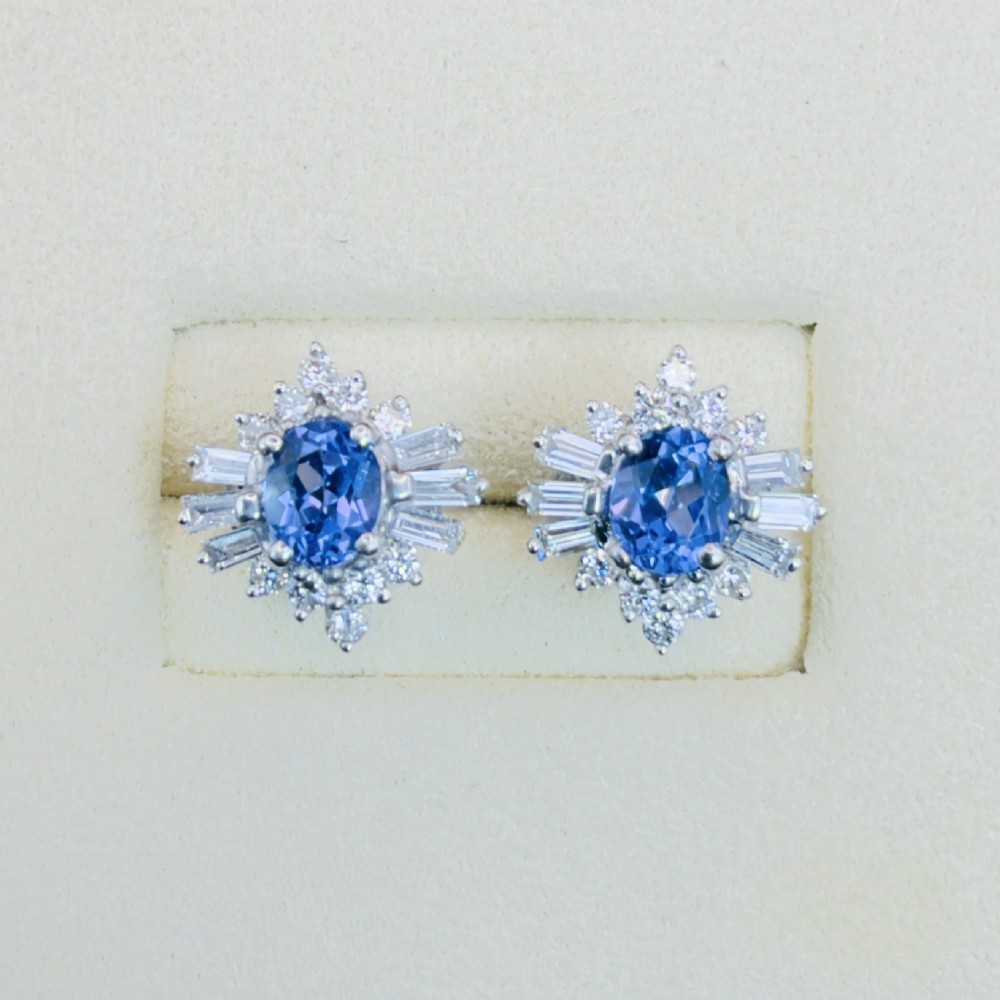 A pair of 18ct white gold cluster earrings featuring central oval cornflower blue sapphires framed by round and baguette-cut diamonds. View II