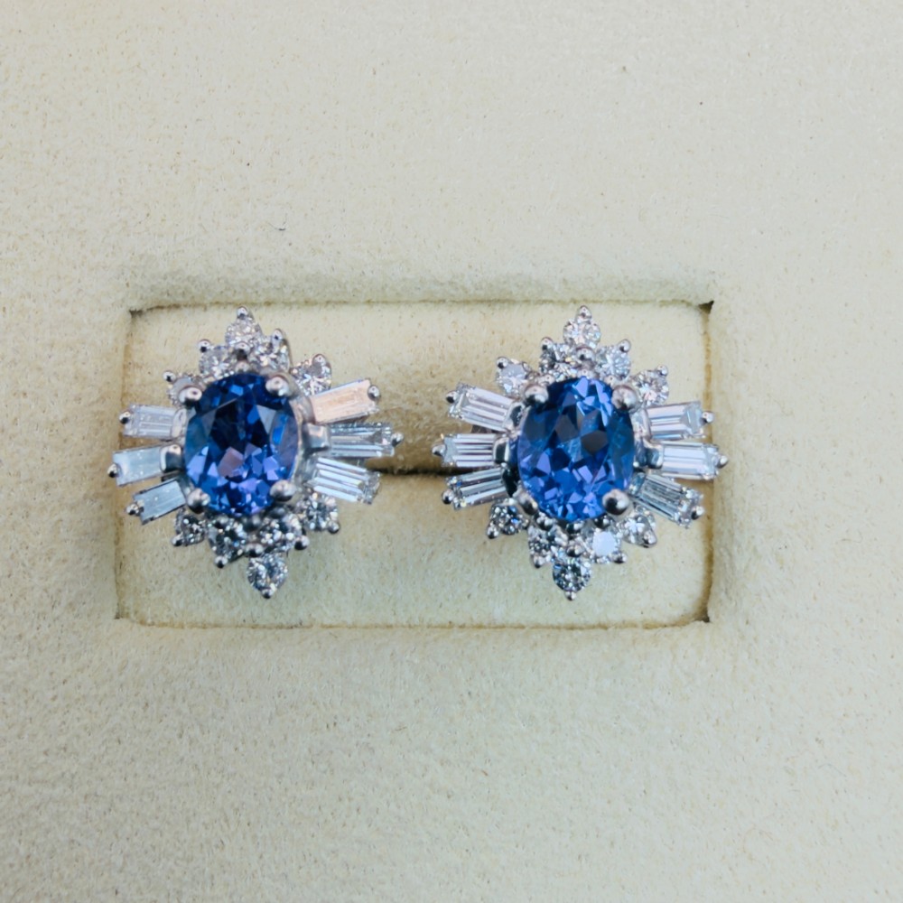 A pair of 18ct white gold cluster earrings featuring central oval cornflower blue sapphires framed by round and baguette-cut diamonds. View V