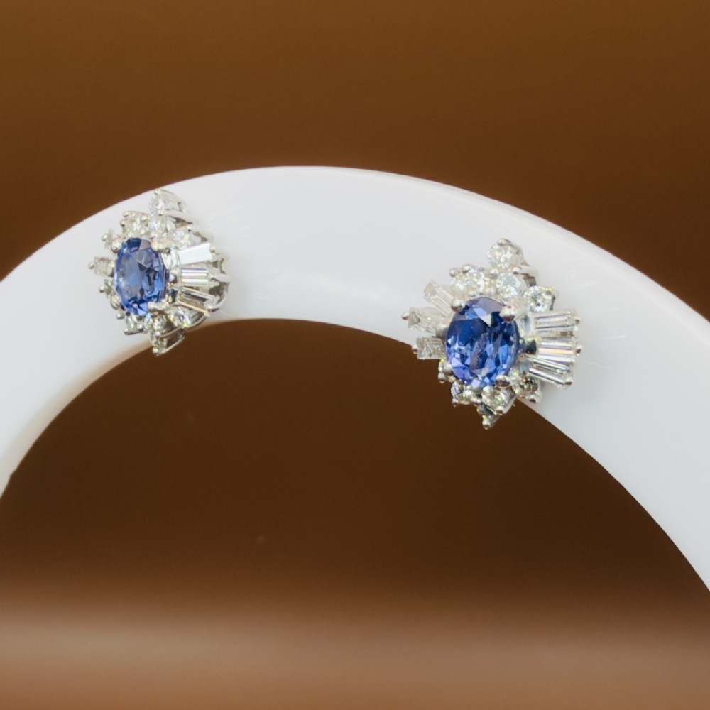 A pair of 18ct white gold cluster earrings featuring central oval cornflower blue sapphires framed by round and baguette-cut diamonds. View IX