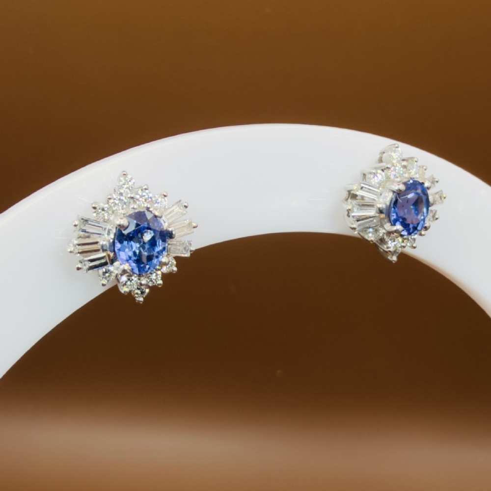 A pair of 18ct white gold cluster earrings featuring central oval cornflower blue sapphires framed by round and baguette-cut diamonds. View X