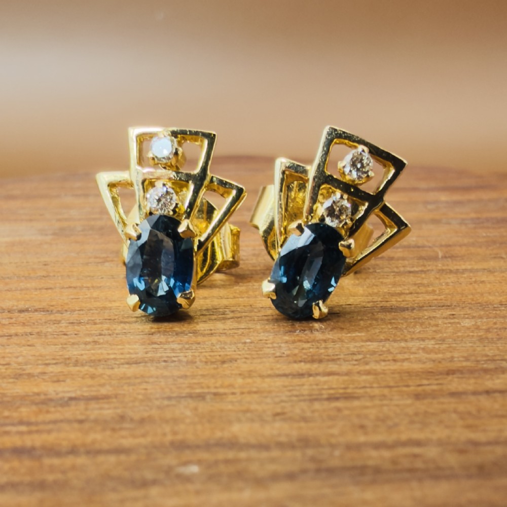 A pair of 18ct yellow gold Art Deco stud earrings featuring an oval blue sapphire topped with two round diamonds inside a geometric openwork frame. View V