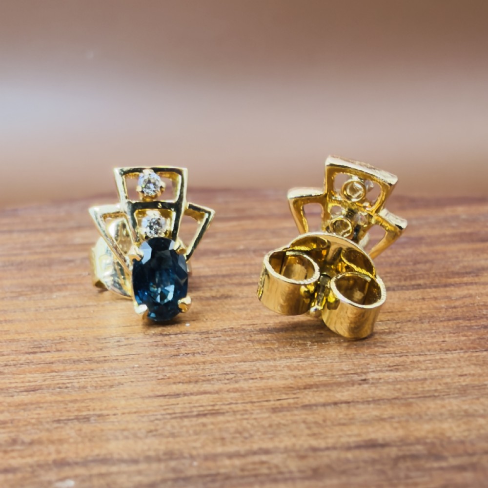 A pair of 18ct yellow gold Art Deco stud earrings featuring an oval blue sapphire topped with two round diamonds inside a geometric openwork frame. View VI