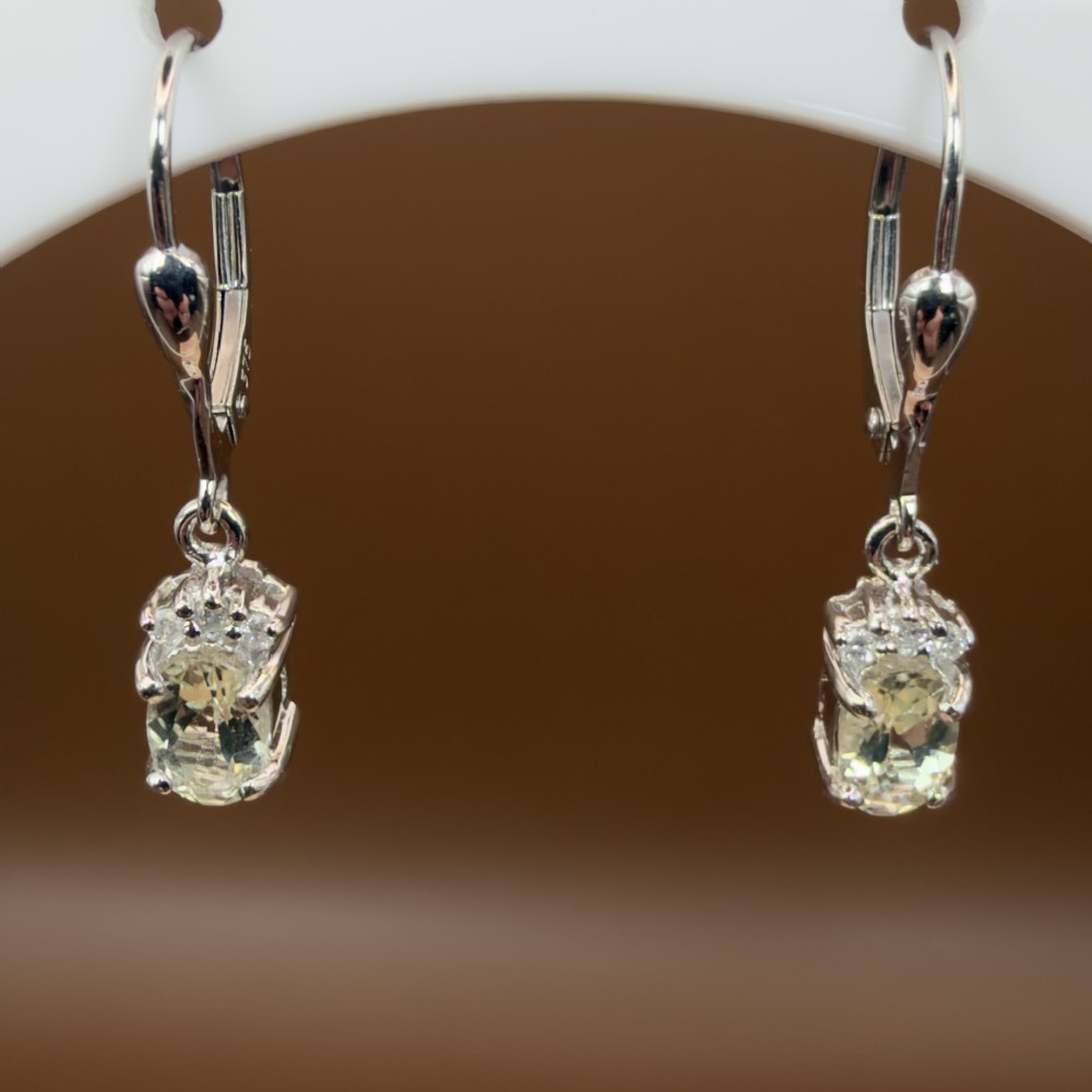 A pair of 9ct white gold drop earrings featuring oval cut pale green amethysts suspended beneath small diamond cluster accents with hinge back fastenings.