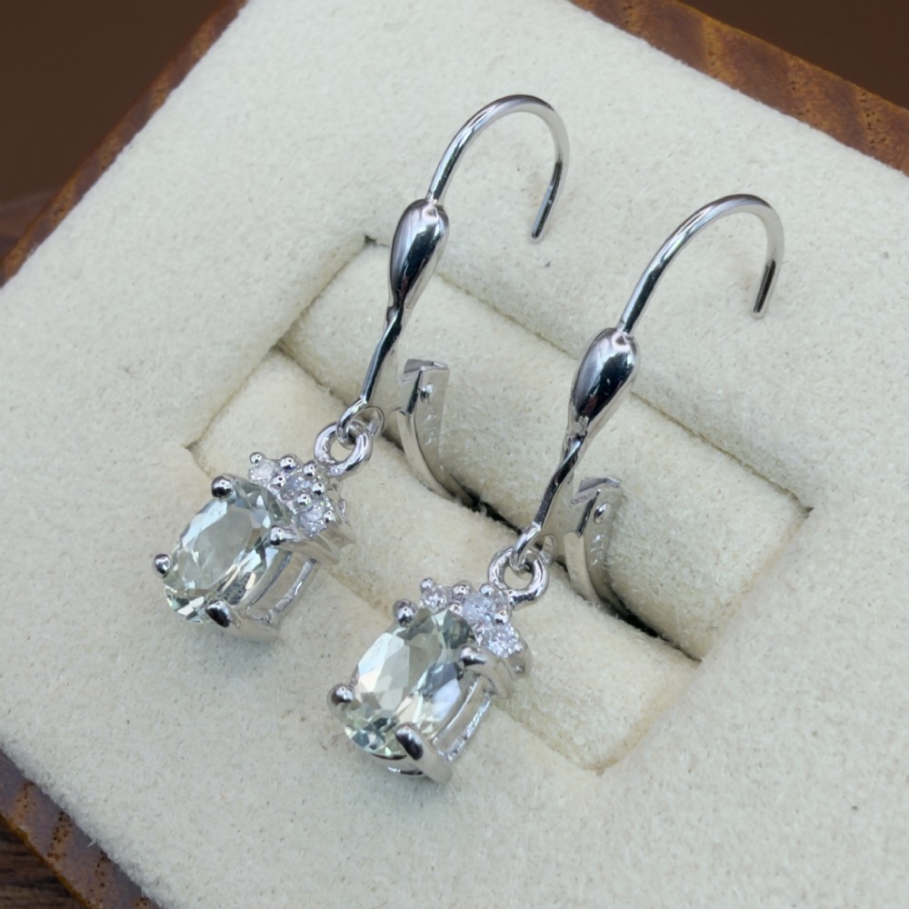 A pair of 9ct white gold drop earrings featuring oval cut pale green amethysts suspended beneath small diamond cluster accents with hinge back fastenings. View VI