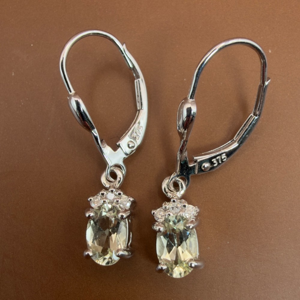 A pair of 9ct white gold drop earrings featuring oval cut pale green amethysts suspended beneath small diamond cluster accents with hinge back fastenings. View IX