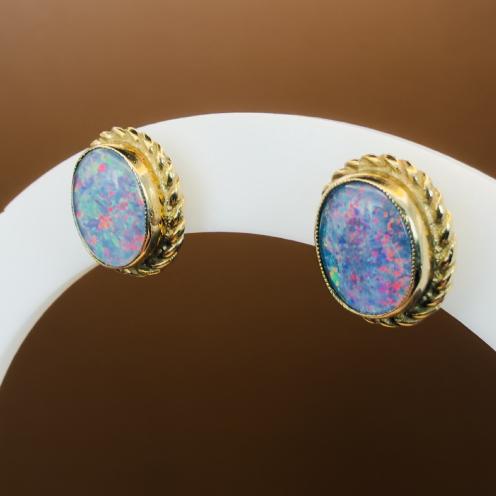 A pair of oval 9ct yellow gold stud earrings featuring iridescent black opal doublets surrounded by a twisted rope border.