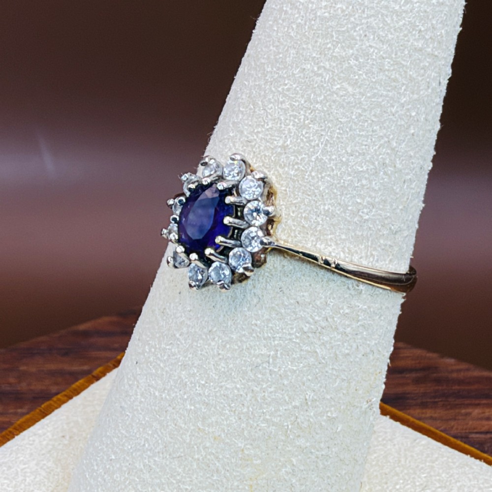 A vintage 9ct yellow gold ring featuring a central oval purple amethyst framed by a halo cluster of round white spinel gemstones. View VI