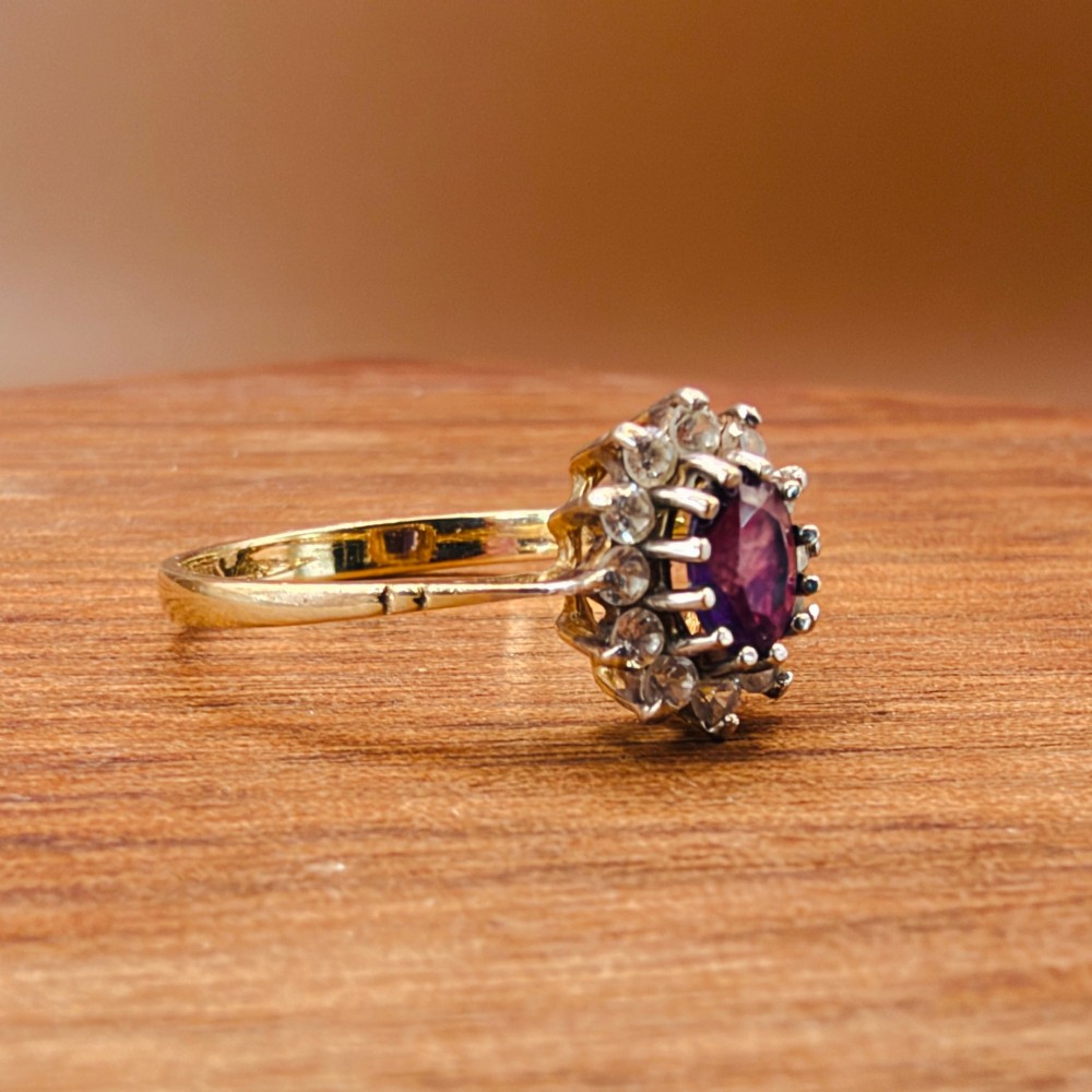 A vintage 9ct yellow gold ring featuring a central oval purple amethyst framed by a halo cluster of round white spinel gemstones. View IX