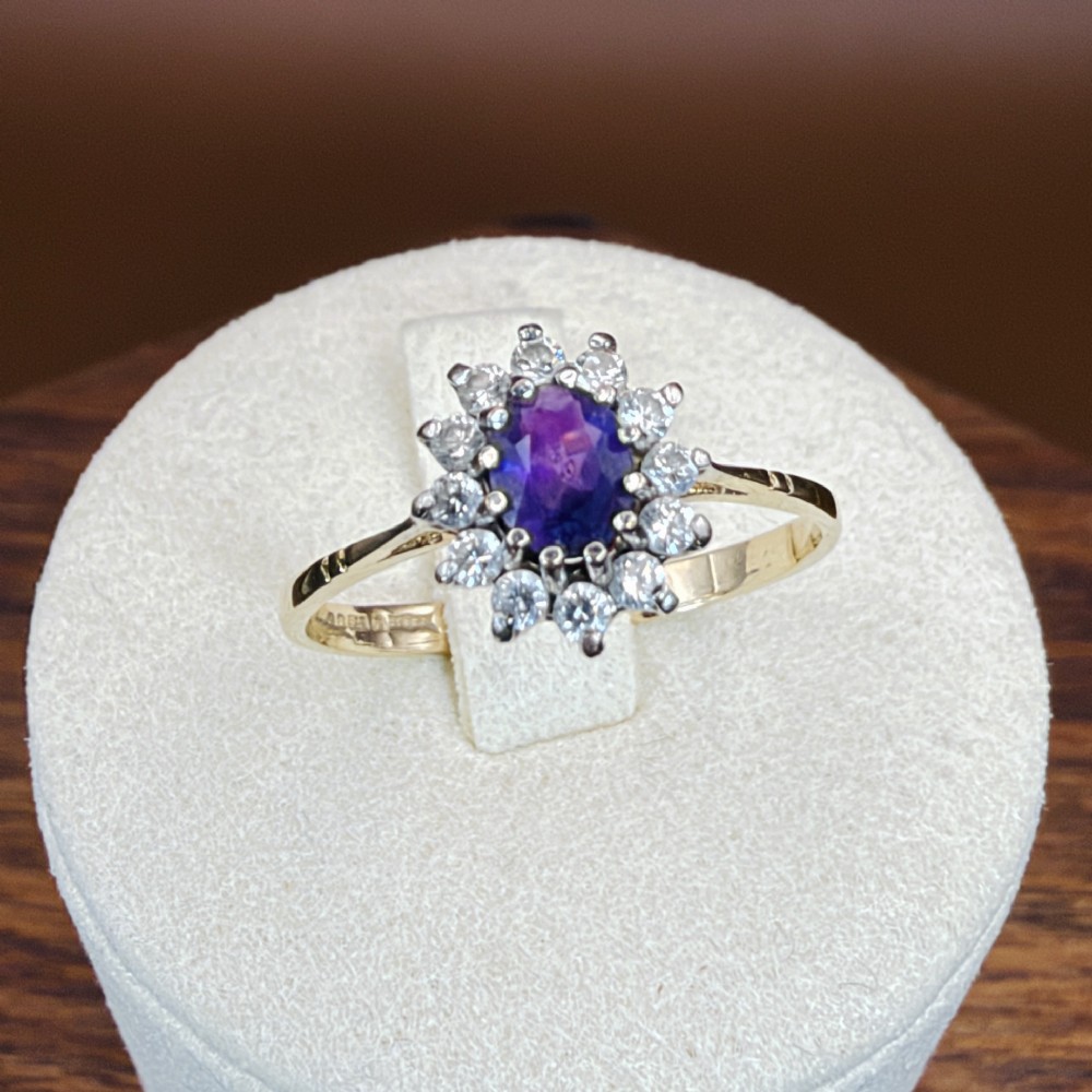 A vintage 9ct yellow gold ring featuring a central oval purple amethyst framed by a halo cluster of round white spinel gemstones. View X