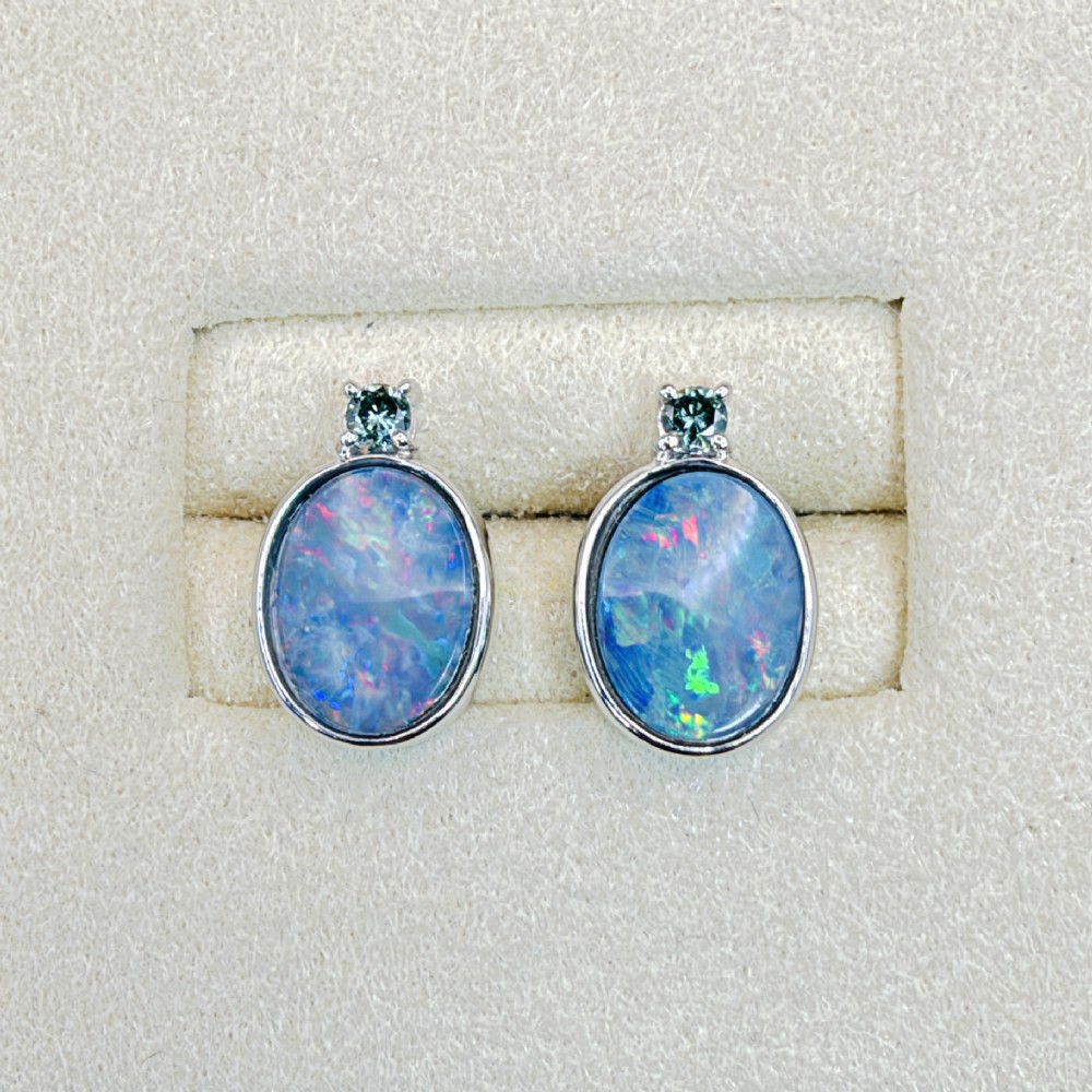 A pair of 14ct white gold stud earrings featuring oval opal doublets with blue and green flashes, each crowned with a small round prong-set teal green diamond. View II