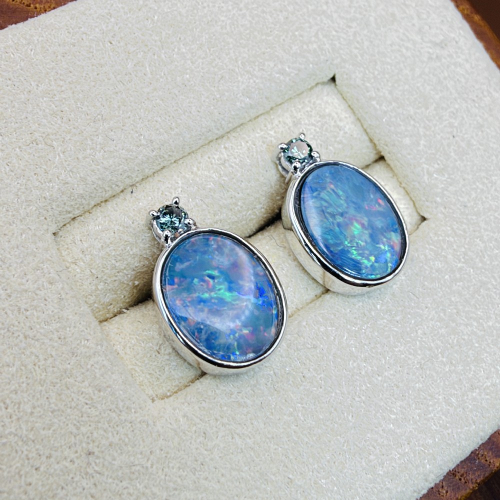 A pair of 14ct white gold stud earrings featuring oval opal doublets with blue and green flashes, each crowned with a small round prong-set teal green diamond. View IV